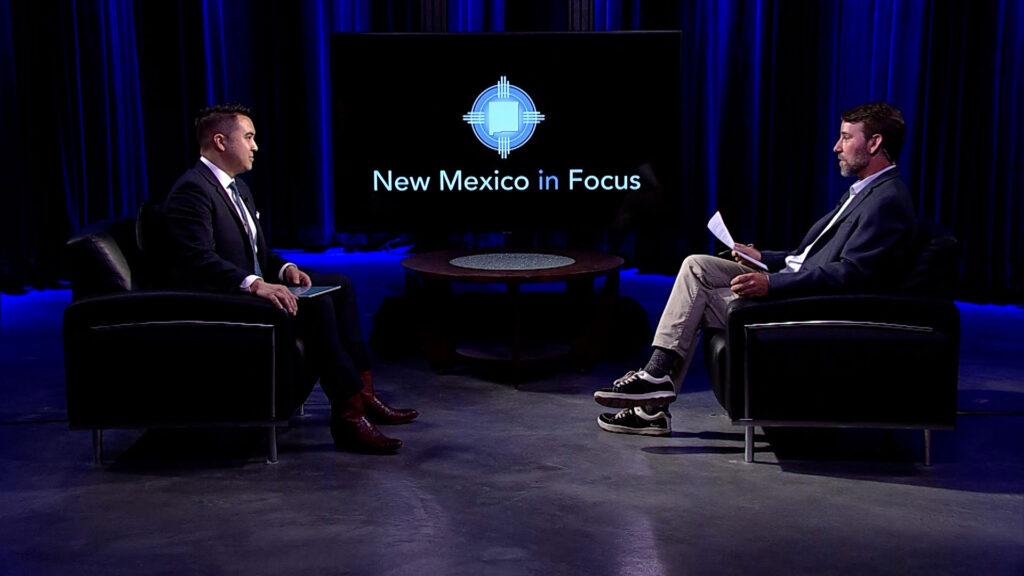 Two men sit facing each other on a TV set with "New Mexico in Focus" displayed on a screen in the background.