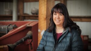 A woman in a dark jacket smiles while standing indoors next to industrial equipment and wooden beams, her eyes reflecting the allure of water.