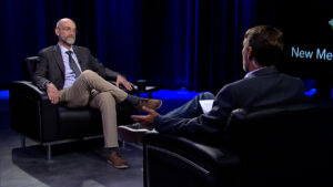 Two men seated in armchairs engaged in a discussion on a dimly lit stage with blue lighting.