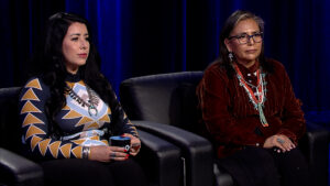 Two women sit on black leather chairs against a dark backdrop. The woman on the left wears a geometric-patterned top, and the woman on the right wears a red shirt with layered necklaces.