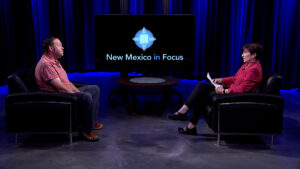 Two people sit facing each other in a studio setting with a "New Mexico in Focus" screen between them.