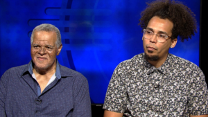 Two men are seated against a blue backdrop, both wearing collared shirts. One has short gray hair, and the other has curly hair and glasses.