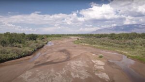 Aerial view of a dry riverbed under a partly cloudy sky, with sparse vegetation and distant mountains in the background.