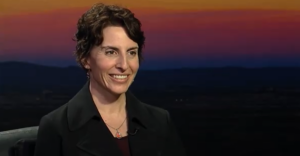 A person with short dark hair smiles, wearing a black coat, against a sunset backdrop.