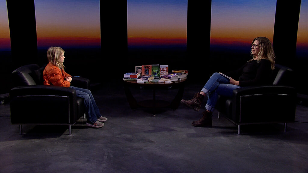 Two people sit in armchairs facing each other on a set with a table of books between them; a backdrop shows a sunset gradient.