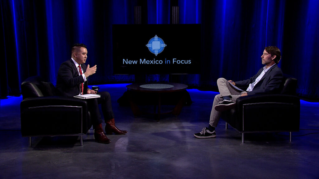 Two men sit facing each other in a studio with blue lighting. The backdrop displays "New Mexico in Focus" on a screen.