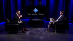 Two men sit facing each other in a studio with blue lighting. The backdrop displays "New Mexico in Focus" on a screen.