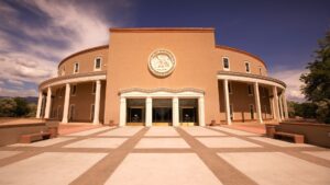 The image shows the New Mexico State Capitol, a round building with columns and the state seal above its entrance, under a blue sky.