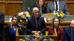 A woman stands at a podium in a legislative chamber, surrounded by seated officials and floral arrangements.