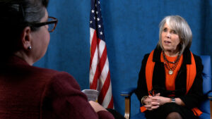 Two women in conversation; one faces away from the camera, and the other is seated in front of a U.S. flag, speaking and gesturing, against a blue backdrop.