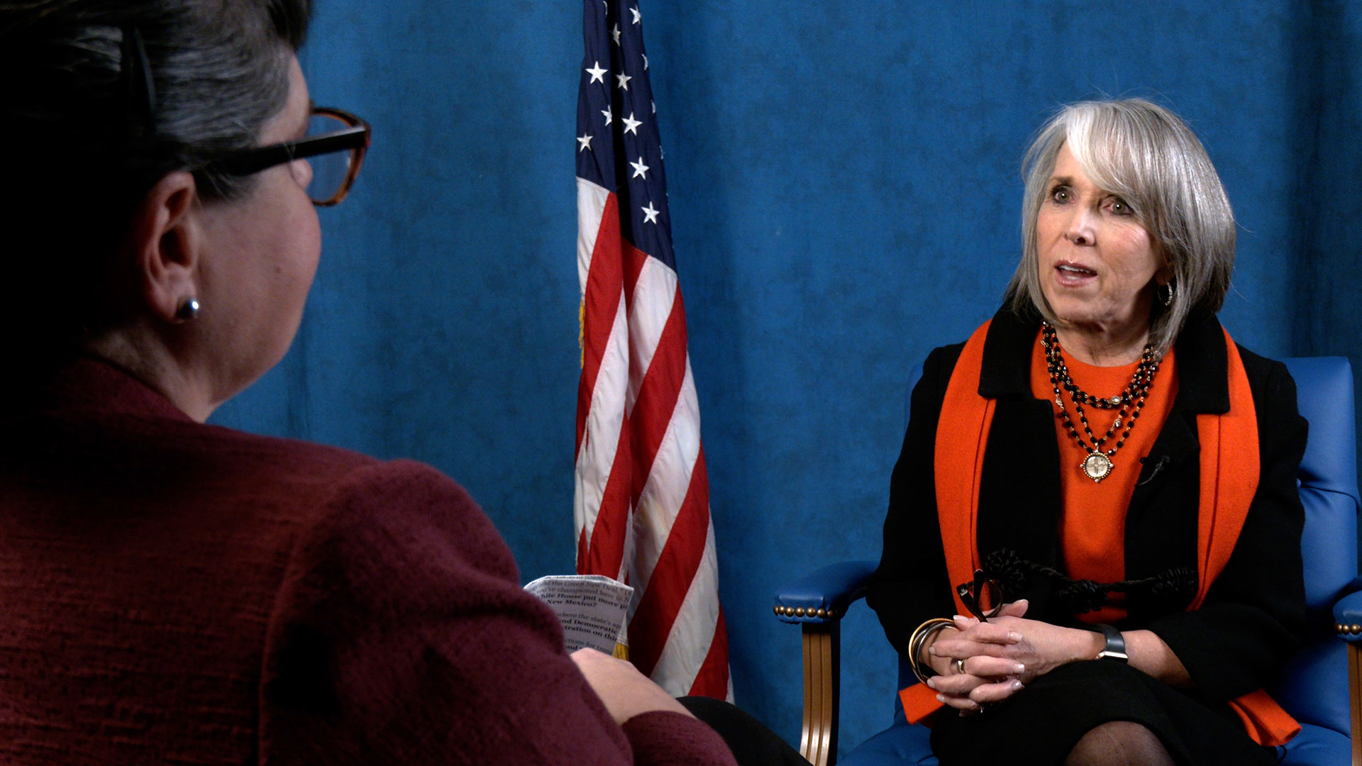 Two women in conversation; one faces away from the camera, and the other is seated in front of a U.S. flag, speaking and gesturing, against a blue backdrop.