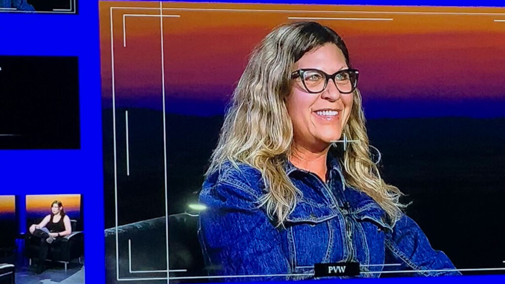 A person with long hair and glasses, wearing a denim jacket, is sitting and smiling against a sunset background, the warm hues reflecting off nearby water.