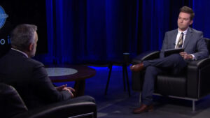 Two men sit facing each other on a dimly lit stage. One is holding a tablet, and they appear to be engaged in a conversation.