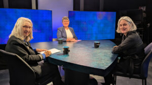 Three women seated around a table in a studio setting with blue screens in the background.