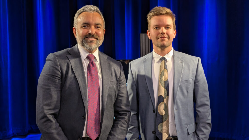 Two men in suits stand side by side in front of blue curtains, both looking at the camera.