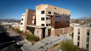 A modern, multi-story beige building with large windows and a central entrance, surrounded by desert landscaping and located on a city street.