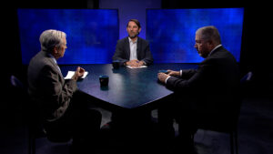 Three men in suits sit around a triangular table, engaged in conversation on a television set with blue screens in the background.