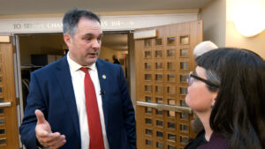 A man in a suit with a red tie talks to a woman wearing glasses and a dark jacket in a hallway with open double wooden doors.