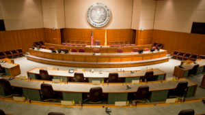 A government legislative chamber with semi-circular seating, desks, and microphones. The room features a large emblem on the wall and an American flag.