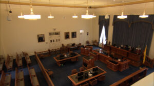 An empty courtroom with wooden desks, chairs, and a judge's bench. Portraits and blue curtains adorn the walls, with computers and lights overhead.