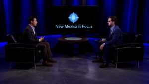 Two people are seated across from each other in a dimly lit studio set with a screen displaying "New Mexico in Focus.