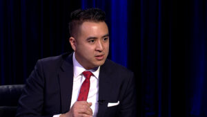 A man in a suit and red tie speaks, gesturing with one hand, against a dark curtain backdrop.