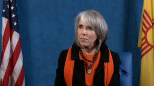 A seated person with gray hair wearing an orange scarf and black jacket is in front of a blue backdrop, with American and yellow flags visible.