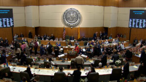 People gather in a legislative chamber with a large seal on the wall, digital boards displaying information, and floral arrangements on desks.