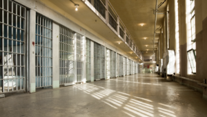 A long hallway with barred prison cells on one side and a balcony above. The floor and walls are concrete, with overhead lighting. Sunlight streams in through high windows, casting shadows.