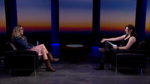 Two people seated in armchairs facing each other in a dimly lit studio with a sunset backdrop, engaging in a conversation.