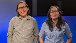 Two people with glasses are smiling and standing side by side in front of a blue and black background.