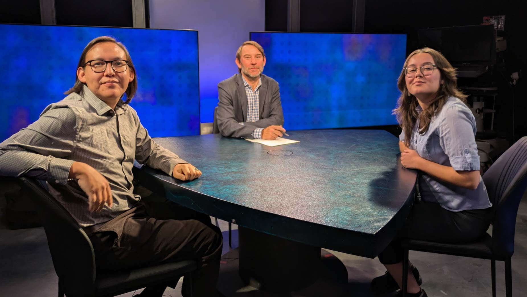 Three people sit around a table in a studio with two large screens behind them. One person holds a piece of paper.