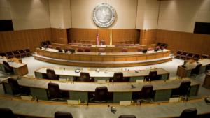 Empty legislative chamber with curved wooden desks, maroon chairs, and a large emblem on the wall. An American flag stands beside the central lectern.