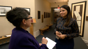 Two women engaging in conversation in a hallway. One holds a notepad, the other a phone. Walls display framed documents and artworks.