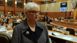 An older woman with short white hair and glasses speaks in a legislative chamber, wearing a black and white polka dot jacket, pearl necklace, and a pin.