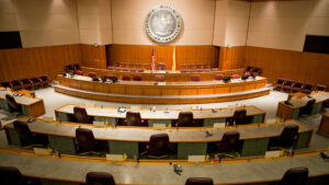 Empty courtroom with a large wooden horseshoe-shaped bench, seal on the wall, and desks with microphones arranged below.