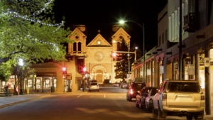 A well-lit street at night leads to a historic church with twin towers, surrounded by parked cars and buildings. Trees with string lights are on the left.