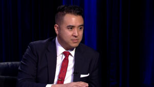 Person in a dark suit and red tie sits in front of a dark curtain, with a focused expression.