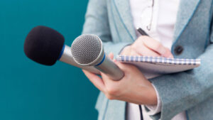 Person in a blue blazer holds two microphones and a notepad, poised to take notes against a teal background.