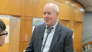 A bald man in a suit and bolo tie is talking to someone off-frame in a wood-paneled room.