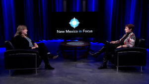 Two people sitting on black chairs facing each other in a studio with blue lighting, discussing on a set labeled "New Mexico in Focus.