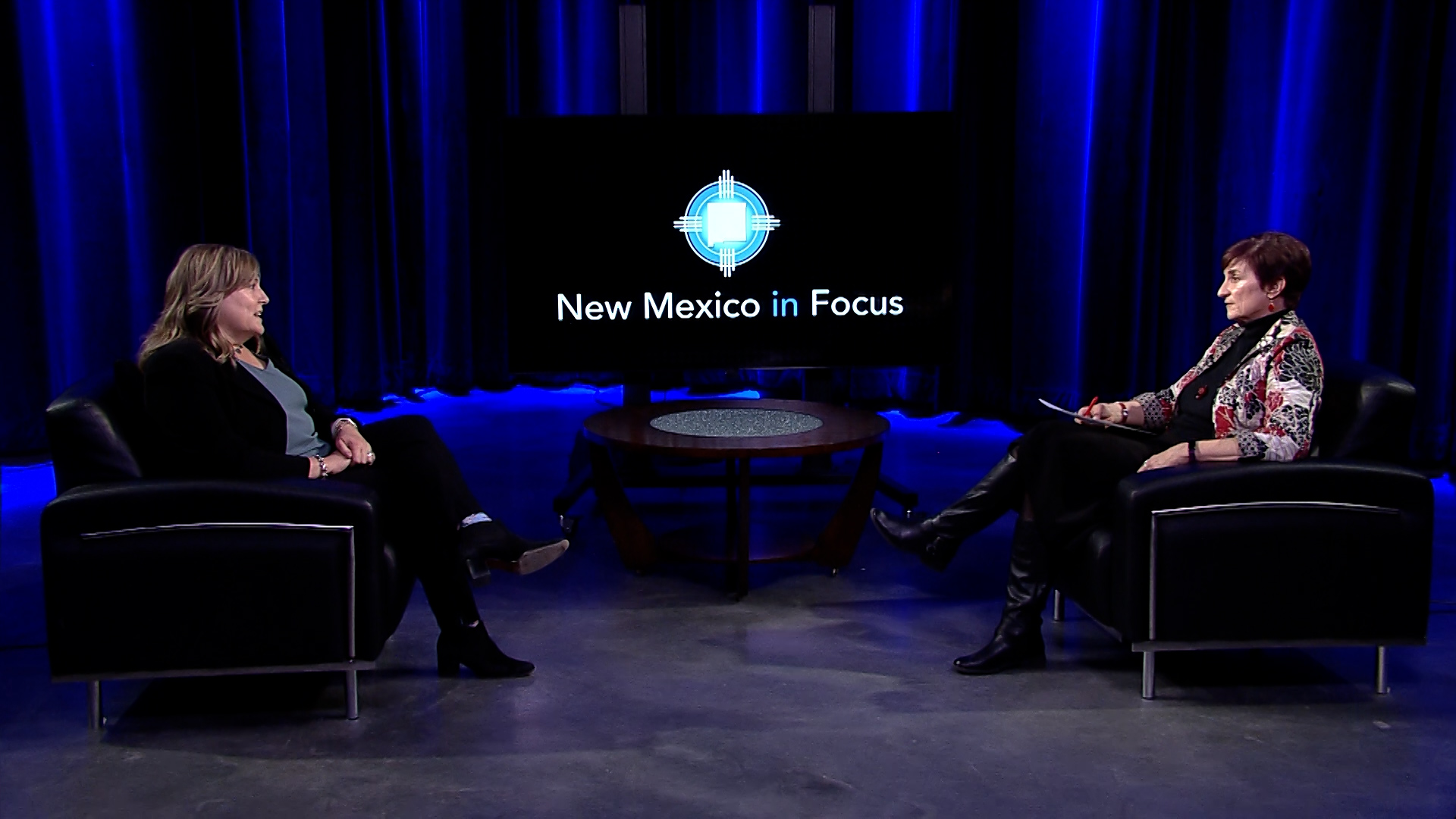 Two people sitting on black chairs facing each other in a studio with blue lighting, discussing on a set labeled "New Mexico in Focus.