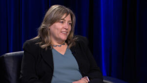 Woman with shoulder-length hair, wearing a black blazer over a teal shirt, sitting and smiling in front of a dark blue curtain.