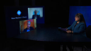 A woman sits at a round table with a tablet, participating in a video conference with two men displayed on large screens. The setup is in a dimly lit studio.