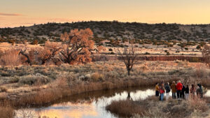 A group of people stands near a reflective pond in a dry, grassy landscape with scattered trees and hills under a clear sky at sunset.