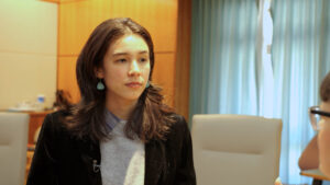 A woman with long brown hair and blue earrings is looking attentively at someone off-camera. She is in a room with wooden walls and blue curtains.