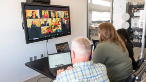 Three people sit at a table in an office, participating in a video conference displayed on a large screen. A laptop and papers are on the table.