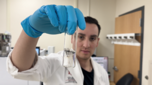 A person in a lab coat and blue gloves holds a small glass container of metallic samples in a laboratory setting.