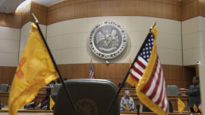 A courtroom with a seal on the wall, flanked by American and New Mexican flags. People are seated in the background.