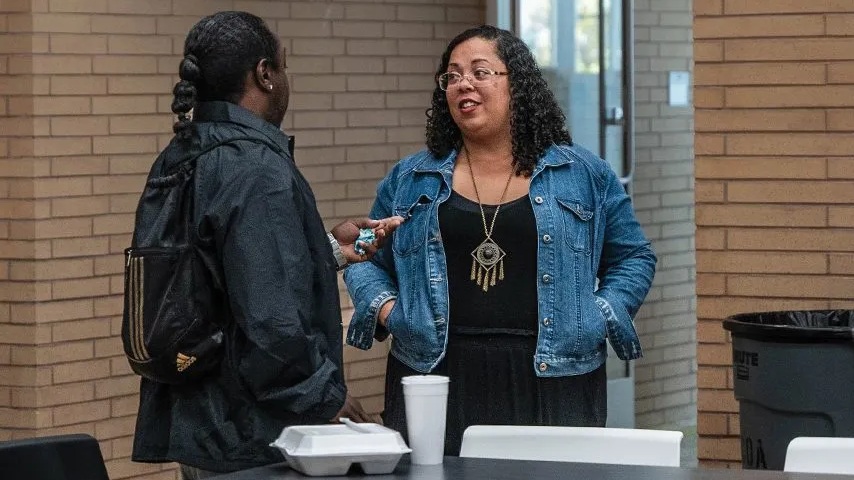 Two people stand and talk indoors near a table with food containers and a cup. One person holds a phone while the other listens. Behind them is a brick wall.